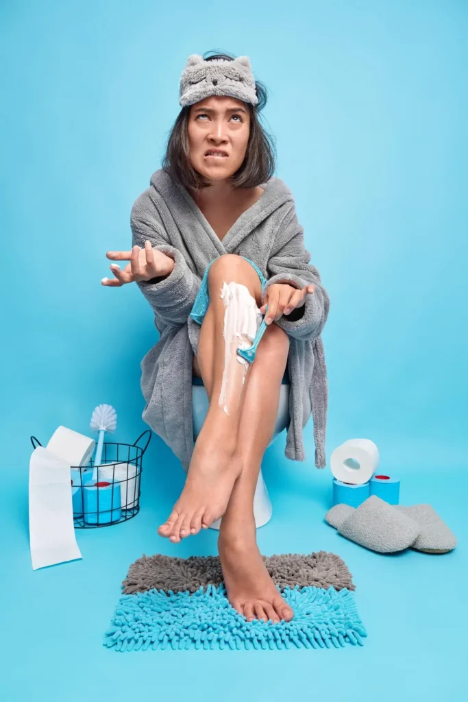 Frustrated woman shaving her legs in a bathroom before a date, wearing a bathrobe and sleep mask — representing improper pre-care habits before laser hair removal that can lead to skin irritation or pigment changes if not avoided