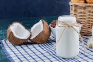 A glass jar of fresh coconut milk beside halved coconuts on a checkered cloth, representing natural and healthy coconut milk.