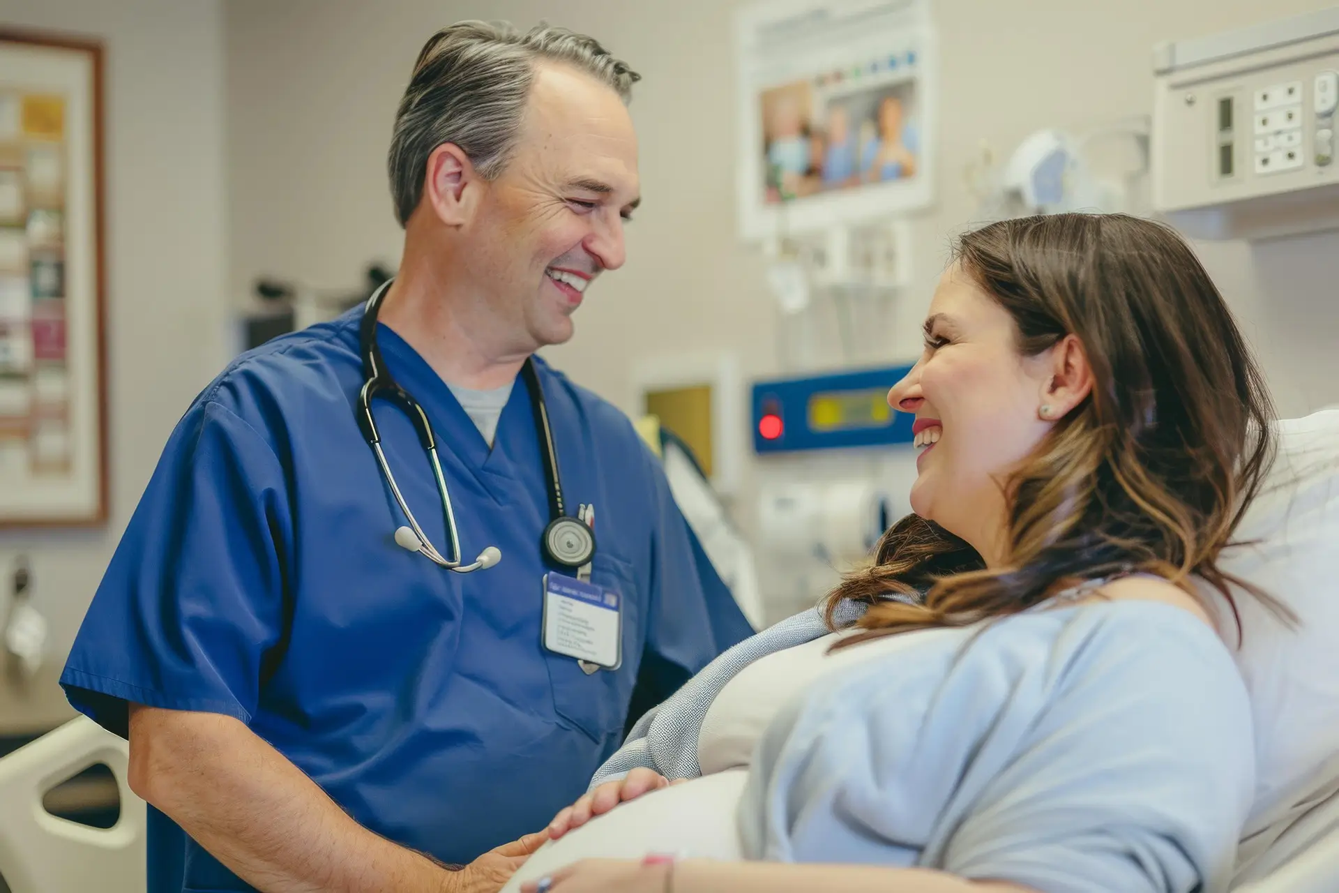 A healthcare professional in blue scrubs smiles while interacting with a pregnant woman in a hospital room, conducting a general physical examination.