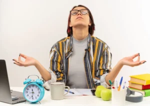 Young woman practicing meditation at her desk to manage stress and stay calm while working, surrounded by books, a laptop, and a cup of coffee.