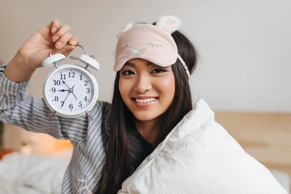 Smiling woman holding an alarm clock and wearing a cute sleep mask — symbolizing good sleep hygiene practices that help prevent mouth breathing at night