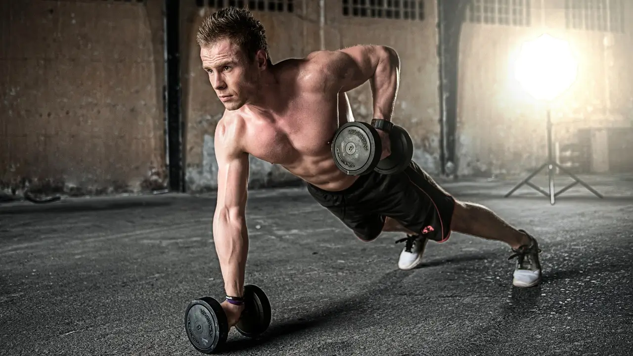A man performing a strength training exercise with dumbbells, engaging in a push-up-like movement for building upper body strength in a gym setting.