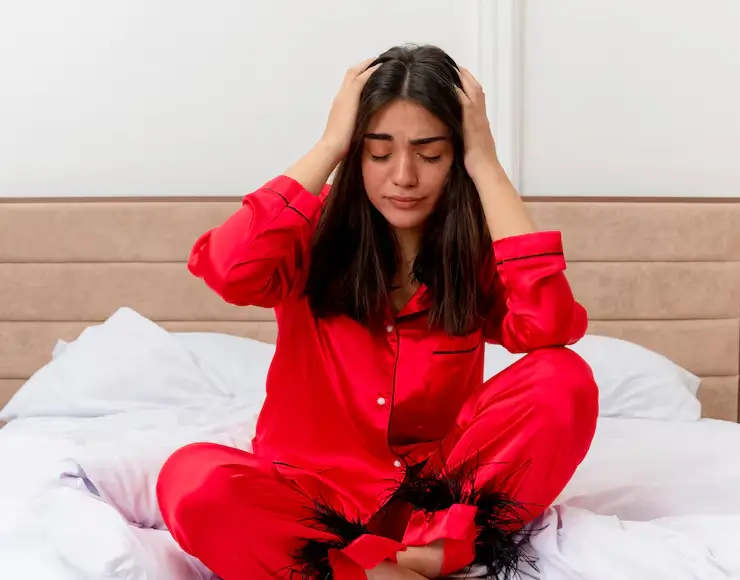 Young woman resting in bed wearing red pajamas, holding her lower abdomen as if experiencing discomfort from a urinary tract infection.