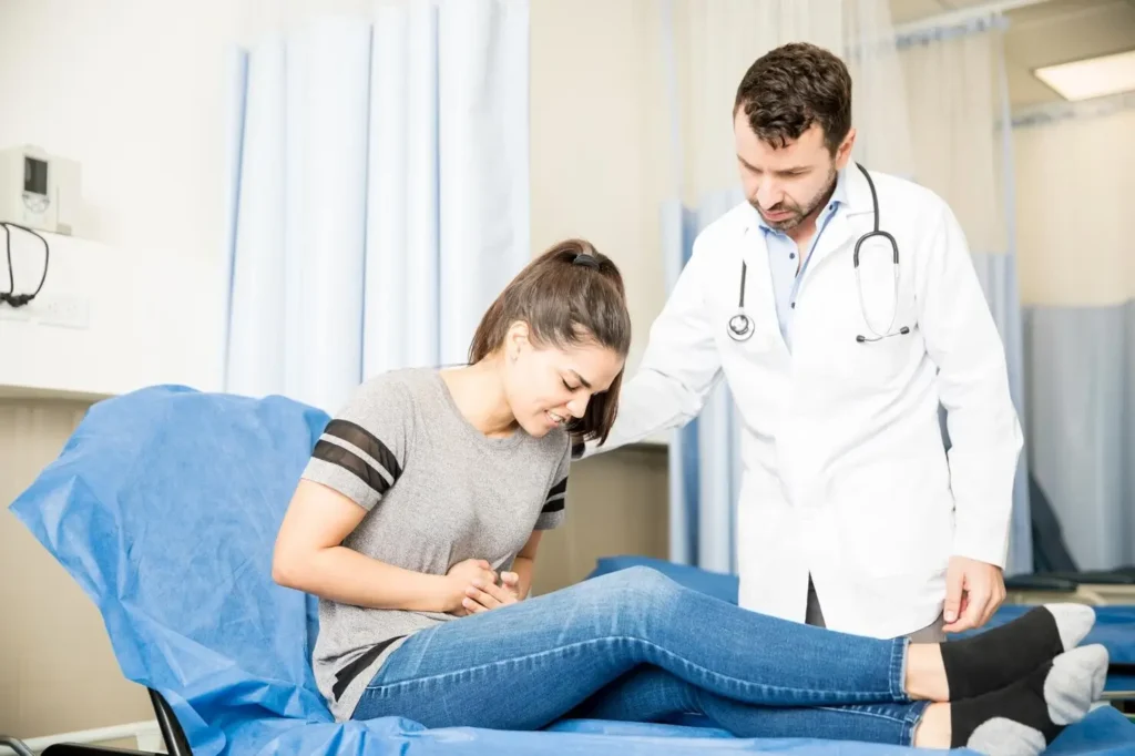 Young woman holding her lower abdomen in pain while a doctor examines her in a medical clinic, suggesting symptoms of a possible urinary tract infection.